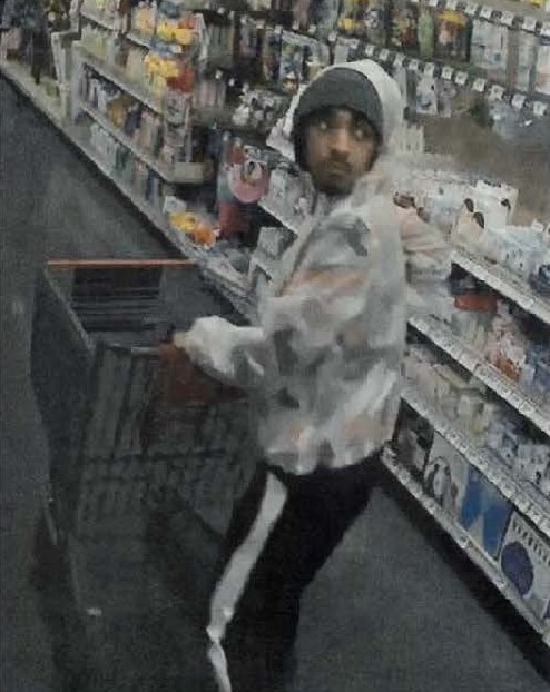 Photo of suspect at HEB pushing a shopping cart, wearing a large gray beanie with a light colored hoodie and black jogging pants with a thick white stripe going down the side.  